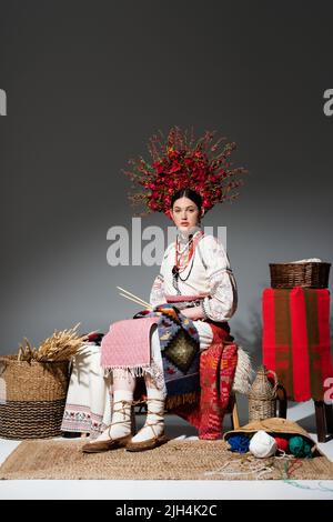 full length of brunette ukrainian woman in traditional clothes and wreath with flowers holding knitting needles on dark grey Stock Photo