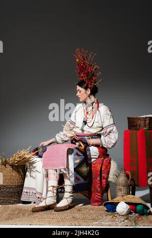 full length of ukrainian woman in traditional clothes and red wreath with flowers holding knitting needles on dark grey Stock Photo