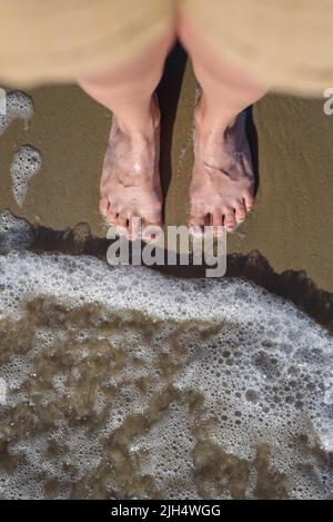 View below to woman barefoot legs from her head standing on sand in wave of Baltic sea Stock Photo