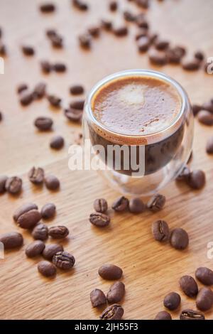 A transparent glass cup of hot espresso standing on a wooden table top, surrounded by coffee beans in the background Stock Photo