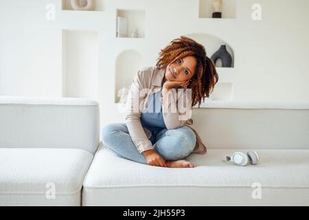 African american millennial girl, contented and happy, sit on comfortable sofa in cozy modern light living room. Enjoy her live Stock Photo