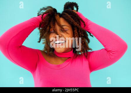 Dreamy young beautiful African American woman with bright eyelines wearing pink bodysuit over blue wall keeps hands pressed together under chin, looks Stock Photo