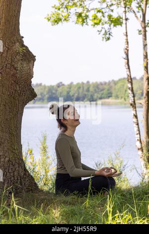 Close-up shots of a young woman practicing yoga in nature. Sit on green grass next to a tree and a forest lake. Communicate to exercise and maintain good health. High quality photo Stock Photo