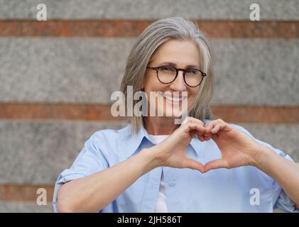Portrait of attractive mature woman with grey hair showing heart sign gesturing with her hands and smile looking at camera wearing blue casual shirt outdoors. Mature people beauty and care concept. Stock Photo