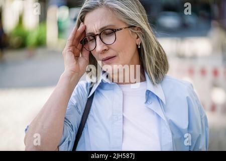 Mature grey haired woman 50s, 60s, standing outdoors holding her head suffering from migraine, headache. Suffering from summer teat feeling sick while travel mature woman standing outdoor. Stock Photo