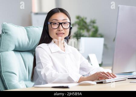 Portrait of a young beautiful Asian woman in headphones with a microphone. Works as a consultant in a call center, answers calls. Sitting at the table, looking at the camera, smiling. Stock Photo