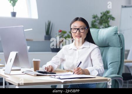 Workaholic. A young Asian business woman, sitting at the table drinking coffee to go, a paper cup of coffee is standing on the desktop near the computer. He looks at the camera, smiles. Stock Photo