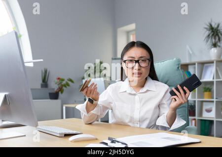 Fraud. Upset young beautiful Asian woman holding phone and credit card. A disappointed young lady lost her life savings to a scam. Sitting at the desk in the office Stock Photo