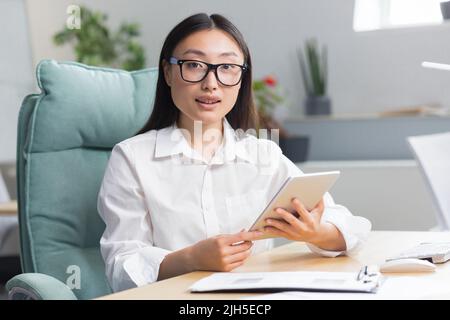 Young beautiful Asian business woman in glasses sits in the office at a computer desk. Holds a tablet in her hands, types, works. Looks at the camera, smiles. Stock Photo