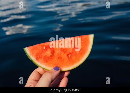 hand holding up watermelon semi circle, half moon with lake, sea, water background on sunny day Stock Photo