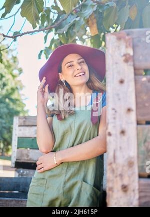 Portrait of a woman in a straw hat standing under a tree next to a rustic wooden crate. One young happy female wearing a summer hat and dungaree dress Stock Photo