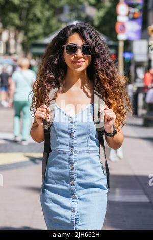attractive dark haired tourist walking the street with a backpack. She wears a denim dress and sunglasses Stock Photo