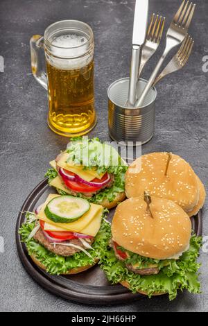 Cheeseburger with greens on round board. Glass mug with beer. Knife and fork in tin. Top view. Black background Stock Photo
