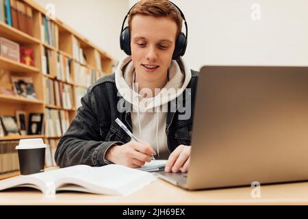 Smiling guy in headphones sitting at desk in library and writing Stock Photo
