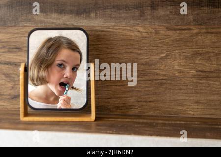 Oral hygiene, healthy teeth and care. Girl brushing teeth with toothbrush and looking in mirror in bathroom interior in the morning, closeup, empty space. High quality photo Stock Photo
