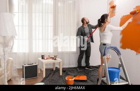 Confident couple painting room with orange color paint to redecorate apartment with paintbrush and roller. Doing diy manual renovation using decorating tools and equipment, housework change. Stock Photo