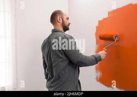 Man renovating apartment with orange paint, using new color on living room wall. Redecorating house with painting tools and paintbrush to change design, home decoration improvement. Stock Photo