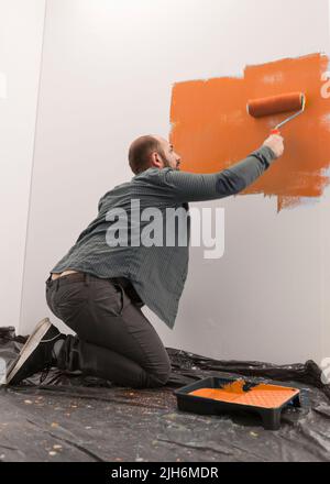 Craftsman using orange paint on room wall, doing handmade painting job with roller and paintbrush. Redecorating apartment with new color, changing house design, housework renovation. Stock Photo