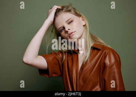 Fashionable stylish woman touching hair posing isolated in brown palette leather dress trench coat isolated on over olive green wall background. Cool Stock Photo