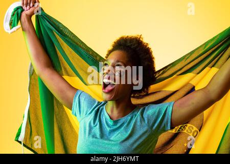 Image of african american female soccer fan with flag of brazil cheering in yellow lighting Stock Photo