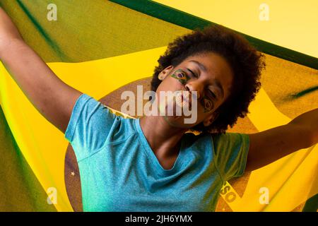 Image of african american female soccer fan with flag of brazil cheering in yellow lighting Stock Photo