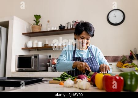 Happy biracial mature woman with short hair wearing apron chopping vegetables on kitchen island Stock Photo