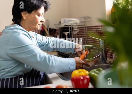 Side view of biracial mature woman washing carrot in colander under running water in kitchen sink Stock Photo