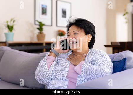 Biracial mature woman with short hair laughing while talking over smartphone and relaxing on sofa Stock Photo