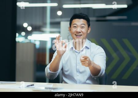 Online webinar. Portrait of an active young Asian businessman. He conducts an online webinar on camera, sits at a table in a modern office, talks, explains, gestures with his hands, smiles. Stock Photo