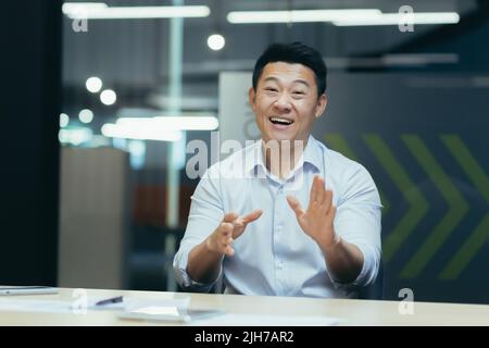 Online webinar. Portrait of an active young Asian businessman. He conducts an online webinar on camera, sits at a table in a modern office, talks, explains, gestures with his hands, smiles. Stock Photo