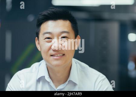 Close-up photo. Portrait of young handsome smiling Asian businessman in white shirt in modern office Stock Photo