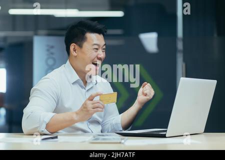 Happy young handsome man Asian businessman rejoices, received income,salary on credit card. He works with a laptop, holds a credit card, sits at a desk in the office Stock Photo