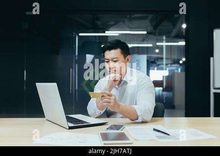 Worried and pensive young man Asian businessman is holding a credit card in his hands, looking at it. Working on a laptop. Sitting in the office at the desk Stock Photo