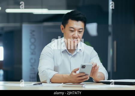 A break at work. A young Asian businessman is using the phone at his desk in a modern office. He holds it in his hands, types a message, makes a phone call. Stock Photo