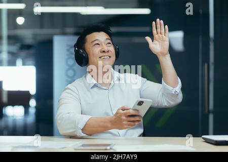 Young man Asian businessman listens to music in headphones and on the phone, took a break from work. He sits in a white shirt in a modern office at a table, dances, makes movements with his hands Stock Photo