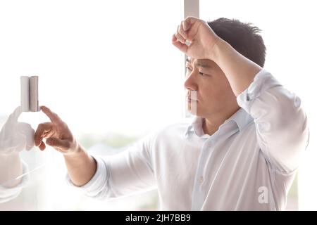 Close-up photo. A young handsome Asian man in a white shirt turns the air conditioner control button on, which hangs on the wall. Standing, leaning against the wall, very hot Stock Photo