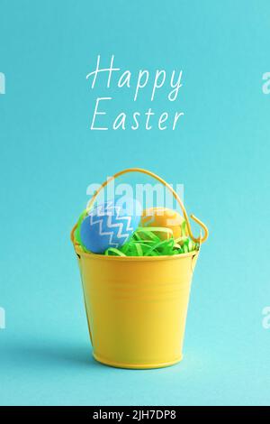 Happy Easter vertical greeting card with decorative colourful eggs in a small yellow bucket isolated on pastel blue background. Stock Photo