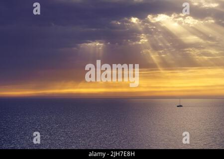 catamaran sailing through the sea with a dramatic sky with solar rays and orange sunset. Spain, Stock Photo