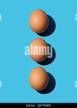 Three brown chicken eggs lie on top of each other on a blue background. Bright light, deep shadow, vertical. Close-up, top view. Stock Photo