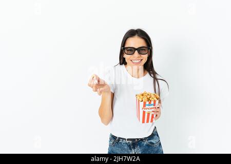 Happy smiling brunette girl in casual jeans and t-shirt wearing 3d glasses eating popcorn, white studio background Stock Photo