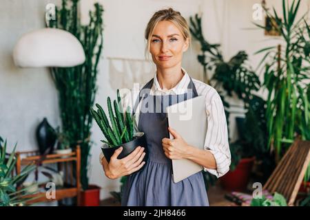 Woman entrepreneur in workshop stands with laptop and potted plant. Stock Photo