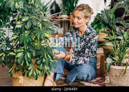 A young woman takes care of potted plants while sitting on the floor in a workshop. Stock Photo
