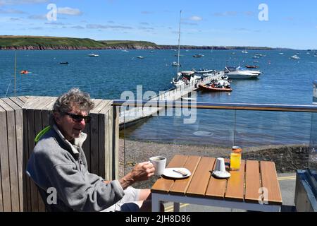 Man sitting having a cup of coffee on an outdoor terrace overlooking Dale Bay, Dale, Pembrokeshire, Wales Stock Photo