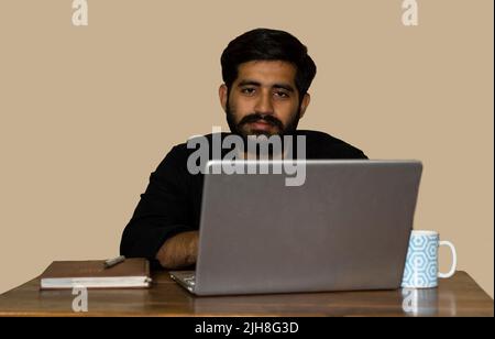 A young Indian man looking at camera while working on laptop. Selective focus on man's face. Stock Photo