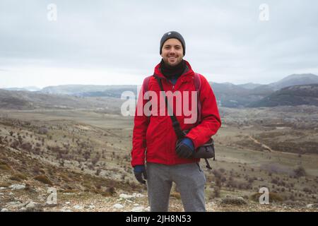A shot of a smiling Caucasian young male wearing a knit cap and red jacket, standing against a landscape Stock Photo