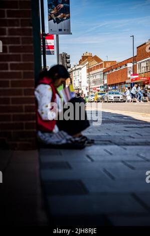 Grantham, Lincolnshire, UK – A young Asian woman sat on the roadside pavement selling copies of The Big Issue as people walk past and ignore her Stock Photo