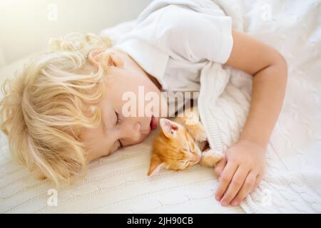 Baby boy sleeping with kitten on white knitted blanket. Child and cat. Kids and pets. Little kid with his animal. Cozy winter evening with pet. Stock Photo
