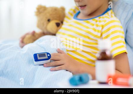 Sick little boy with pulse oximeter on his finger. Asthma treatment. Ill child lying in bed. Unwell kid with chamber inhaler, cough medicine. Stock Photo