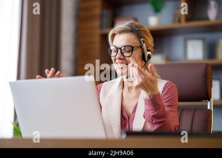 Smiling businesswoman wearing headset talking to laptop web camera sitting in office. Recording vlog, online video conference, video call, online Stock Photo
