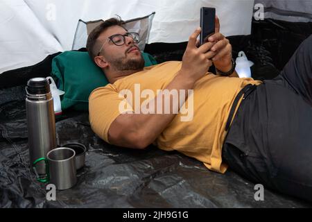 Young adult in yellow t-shirt with glasses lying inside his tent relaxing holding and chatting on his cell phone. Stock Photo